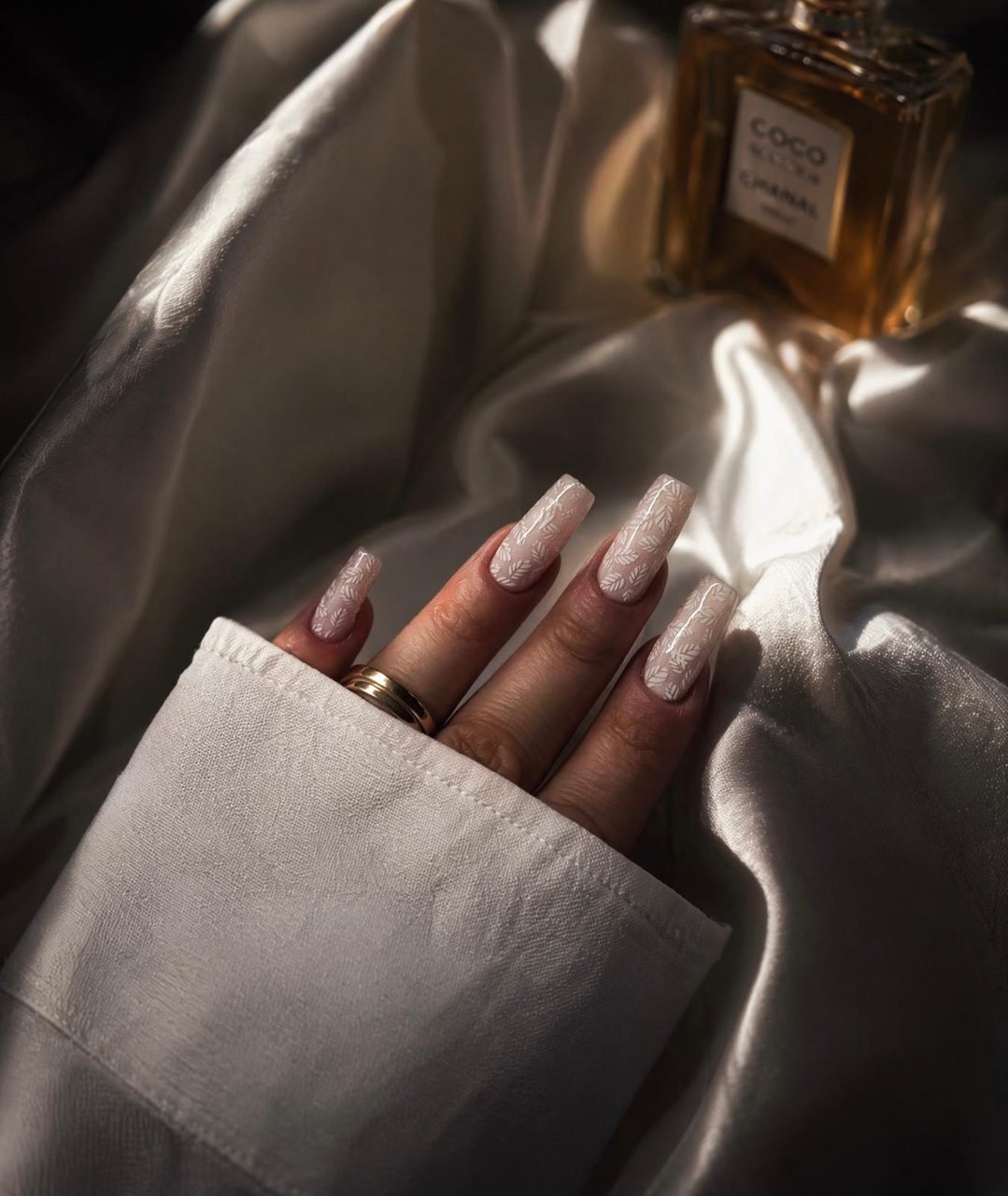 Lange, eckige Nägel in milchigem Nude mit feinem weißem Blätter-Design neben Chanel-Parfum – Nageldesign von Gresa Nails Stuttgart